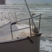 Bow of a beached sailboat on the shore with waves and distant boats in the background.