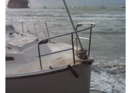 Bow of a beached sailboat on the shore with waves and distant boats in the background.