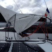 Sailboat's boom with a reefed mainsail, solar panels, and French flag, anchored near a rocky coastline under cloudy skies.