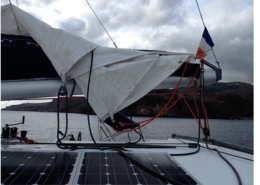 Sailboat's boom with a reefed mainsail, solar panels, and French flag, anchored near a rocky coastline under cloudy skies.