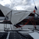 Sailboat's boom with a reefed mainsail, solar panels, and French flag, anchored near a rocky coastline under cloudy skies.