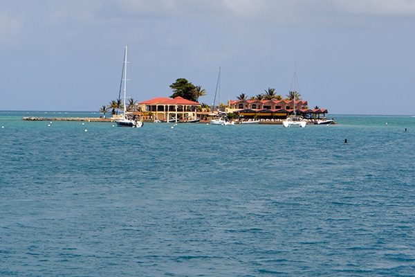 Scenic Caribbean island with sailboats anchored nearby, surrounded by turquoise waters and clear blue sky.