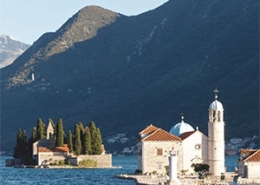 Islands with historic buildings on a calm sea, surrounded by majestic mountains under a clear blue sky.