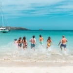 Benefits of flotilla sailing - a group of friends in their bathing suits on the beach running into the water for a swim with sailboats in the background