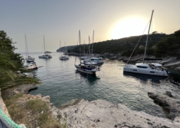 Sailboats anchored in a serene cove at sunset, surrounded by lush hills and a tranquil sea.