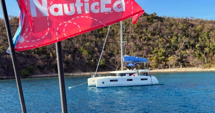 Catamaran sailing near a scenic shoreline, with a red NauticEd flag fluttering in the breeze.