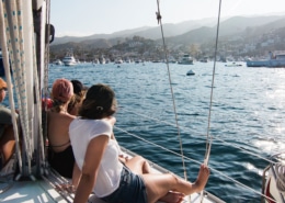 A group of friends enjoying a scenic sail in a sunny bay with boats and hills in the background.