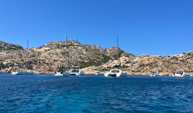Sailboats anchored in clear blue waters near rocky cliffs under a bright blue sky, ideal for Mediterranean sailing adventures.