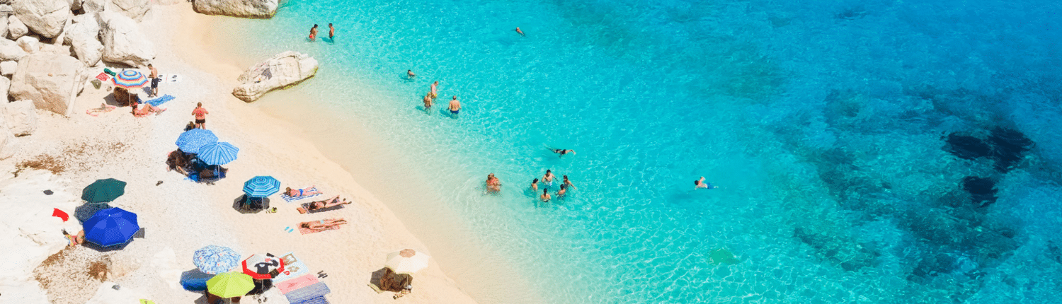 Turquoise sea and sandy beach with colorful umbrellas, rocky cliffs, and sunbathers enjoying a sunny day.