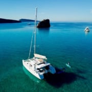 Catamaran gliding on serene blue waters near rocky islet, paddleboarder enjoying the calm sea.