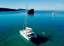 Catamaran gliding on serene blue waters near rocky islet, paddleboarder enjoying the calm sea.