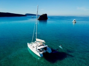 Catamaran gliding on serene blue waters near rocky islet, paddleboarder enjoying the calm sea.
