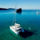 Catamaran gliding on serene blue waters near rocky islet, paddleboarder enjoying the calm sea.