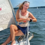 Woman enjoying sailing on a sunny day, seated on a yacht with sails up, surrounded by blue waters and distant shores.