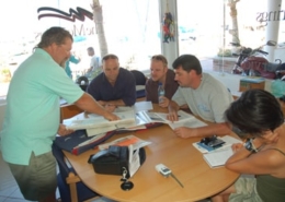 Group of sailors examining navigation charts at a table in a nautical classroom setting.