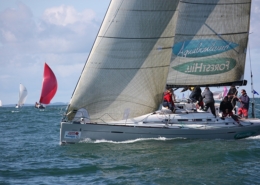 Sailboat racing on open water with crew adjusting sails, vibrant spinnakers in the distance, under a clear sky.