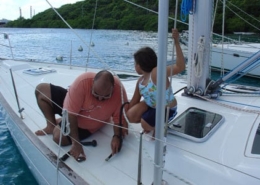 Father and child repairing sailboat deck on a sunny day near lush green shorelines.