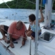 Father and child repairing sailboat deck on a sunny day near lush green shorelines.