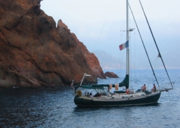 Sailing yacht anchored near rocky cliffs, flag flying high, with calm ocean waters and serene coastal scenery.