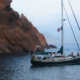 Sailing yacht anchored near rocky cliffs, flag flying high, with calm ocean waters and serene coastal scenery.
