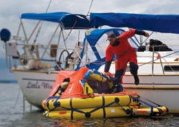 Sailor in survival suit boarding liferaft from yacht for safety drill on calm water, emergency preparedness at sea.