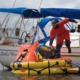 Sailor in survival suit boarding liferaft from yacht for safety drill on calm water, emergency preparedness at sea.