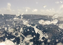 Sea spray splashes over a vast ocean surface under a clear sky, capturing the excitement of open water sailing adventure.