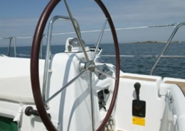 View of a sailboat's helm with a wooden wheel against a clear blue sky and open sea, conveying serene sailing moments.