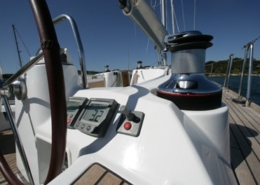 Close-up of a sailboat cockpit with winch, steering wheel, and navigation instruments under a clear blue sky.