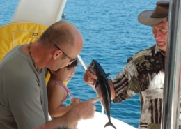 Man showing fish to child and another person on a boat, enjoying a sunny day at sea.