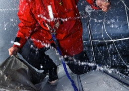 Sailor in a red jacket braving rough seas, harnessed on a wet deck with splashing waves, focused on handling the sail.