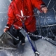 Sailor in a red jacket braving rough seas, harnessed on a wet deck with splashing waves, focused on handling the sail.