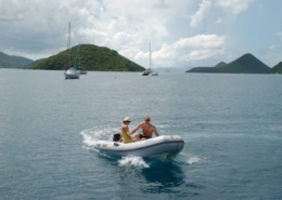 Two people in a dinghy on a calm sea with sailboats and green islands in the background under a cloudy sky.