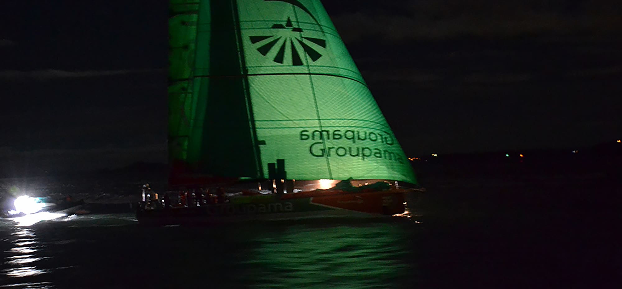 Sailboat Groupama racing at night with illuminated green sail on dark water.