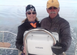 Captain Waddell sailing on a calm sea, smiling at the helm of their boat under a clear sky.