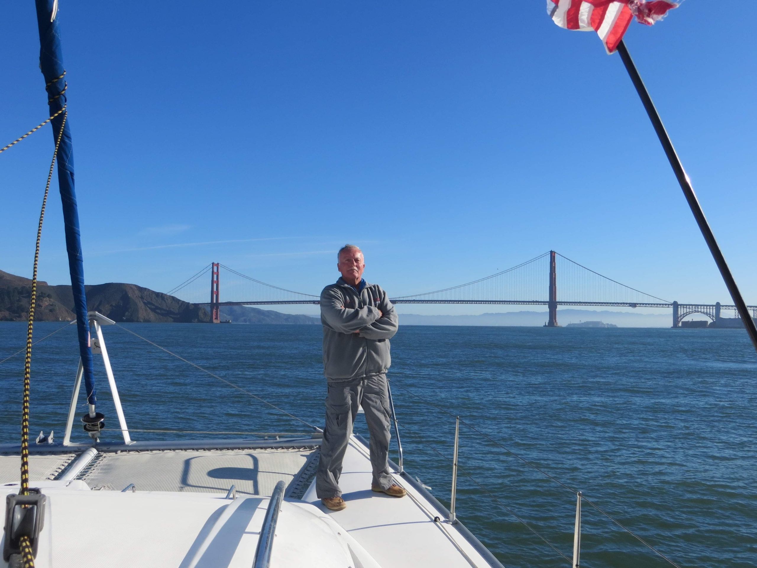 Man standing on a sailboat near Golden Gate Bridge under a clear blue sky.