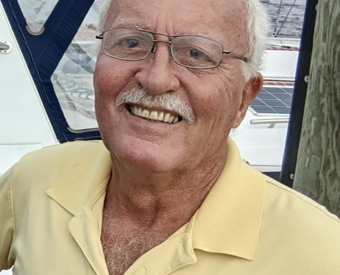 Smiling man on a sailboat by the water, wearing a yellow polo shirt, enjoying a day of sailing.