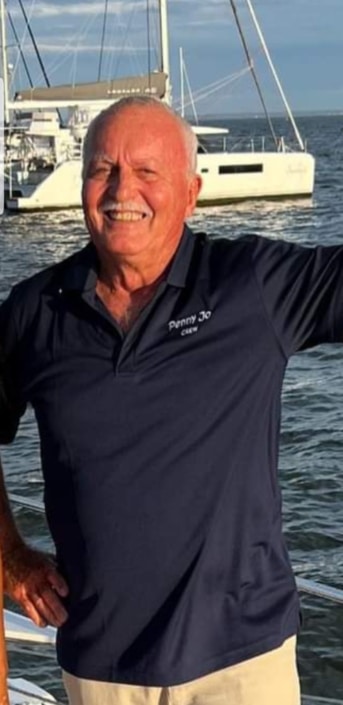 Man smiling on a sailboat wearing a Penny Jo Crew shirt with catamaran in the ocean background.