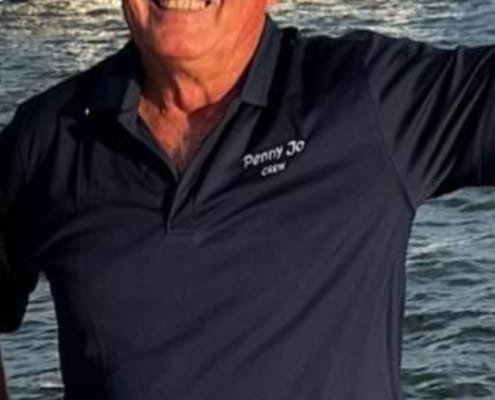 Man smiling on a sailboat wearing a Penny Jo Crew shirt with catamaran in the ocean background.