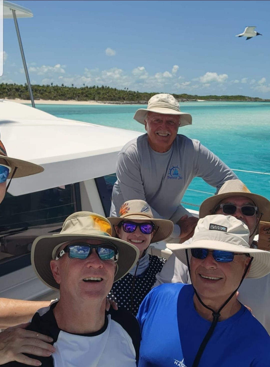 Smiling group sailing on a sunny day in turquoise waters, wearing hats and sunglasses, with a seagull in the sky.