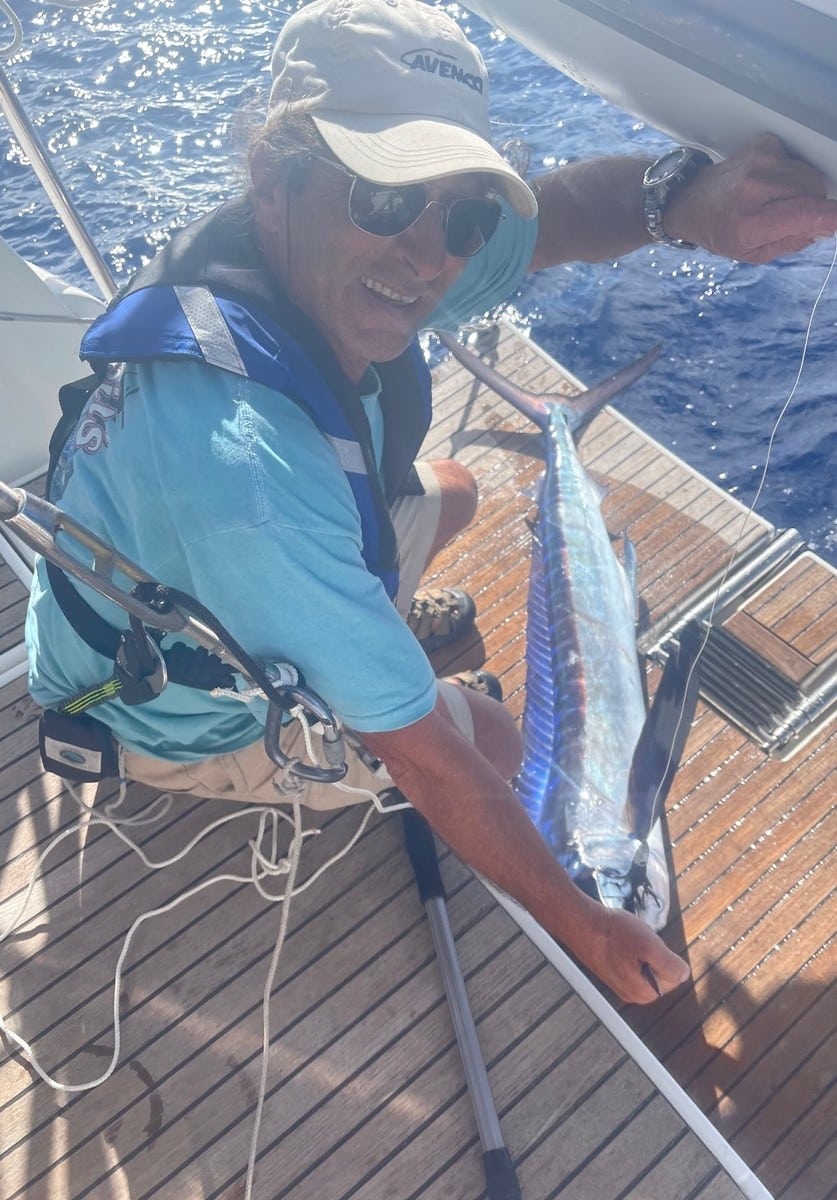 Sailor catches marlin on yacht deck with ocean backdrop, wearing a life vest and cap.