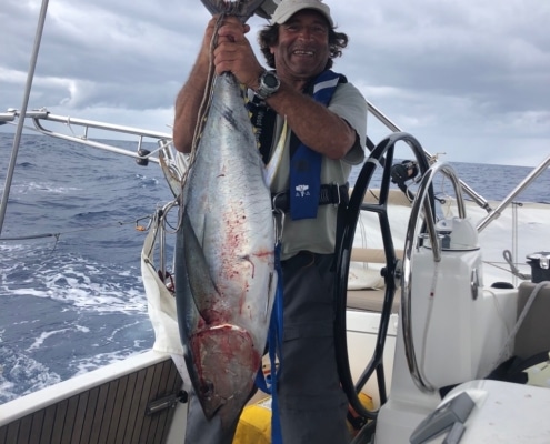 Man proudly displays large tuna catch on a sailboat, ocean in the background, under a cloudy sky.