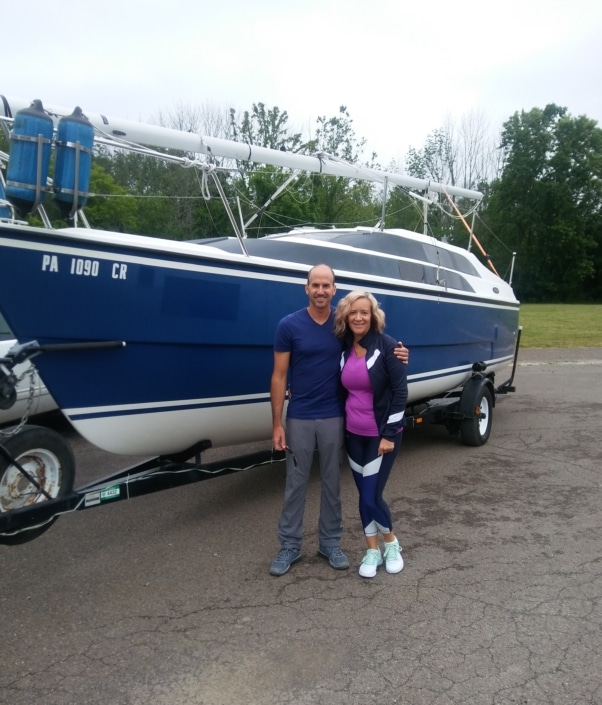 Couple smiling in front of a parked blue sailboat on a trailer, ready for a day of sailing adventure.