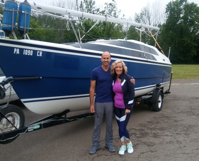 Couple smiling in front of a parked blue sailboat on a trailer, ready for a day of sailing adventure.