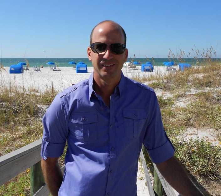 Man in sunglasses on a beach walkway with blue umbrellas and sea in the background on a sunny day.