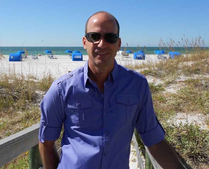 Man in sunglasses on a beach walkway with blue umbrellas and sea in the background on a sunny day.