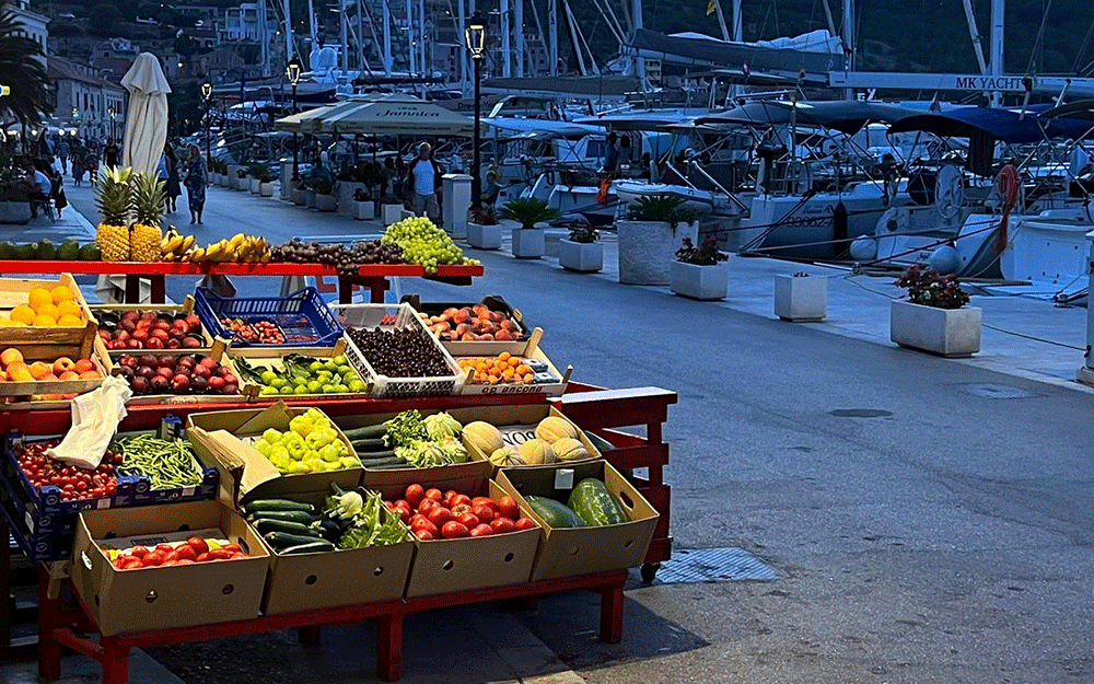 Fruit stand by a marina with various boats docked, showcasing vibrant produce near the waterfront at dusk.