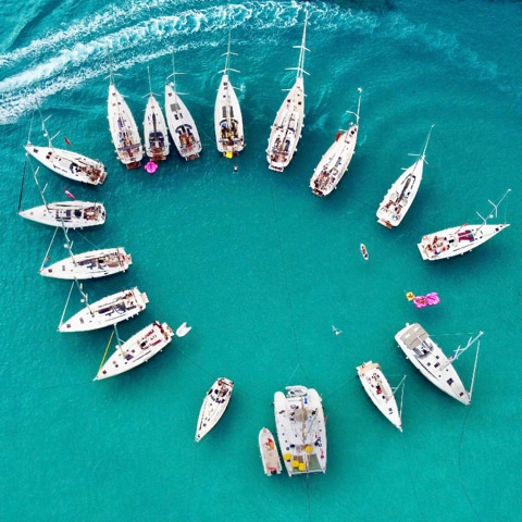 Aerial view of sailboats arranged in a circular formation on turquoise waters, creating a unique social gathering at sea.