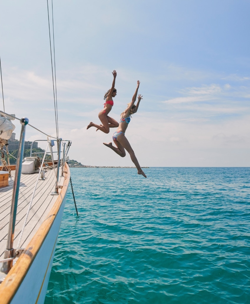 Two women joyfully jump off a sailboat into the blue sea under a clear sky, capturing the spirit of summer adventure.