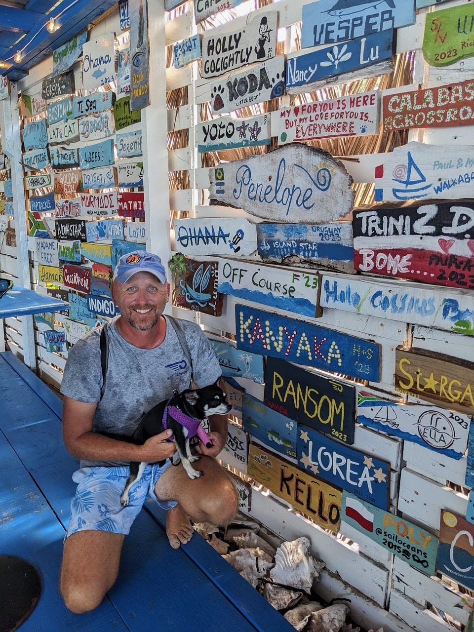 Man with a dog kneels by a colorful wall of boat name plaques at a coastal marina, surrounded by vibrant sailing signage.